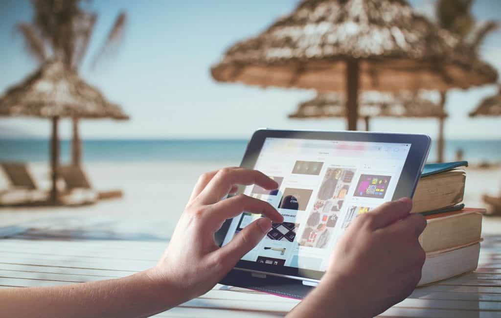 Close up of person's hands holding and scrolling electronic tablet with tropical beach and sun loungers/shades in the background
