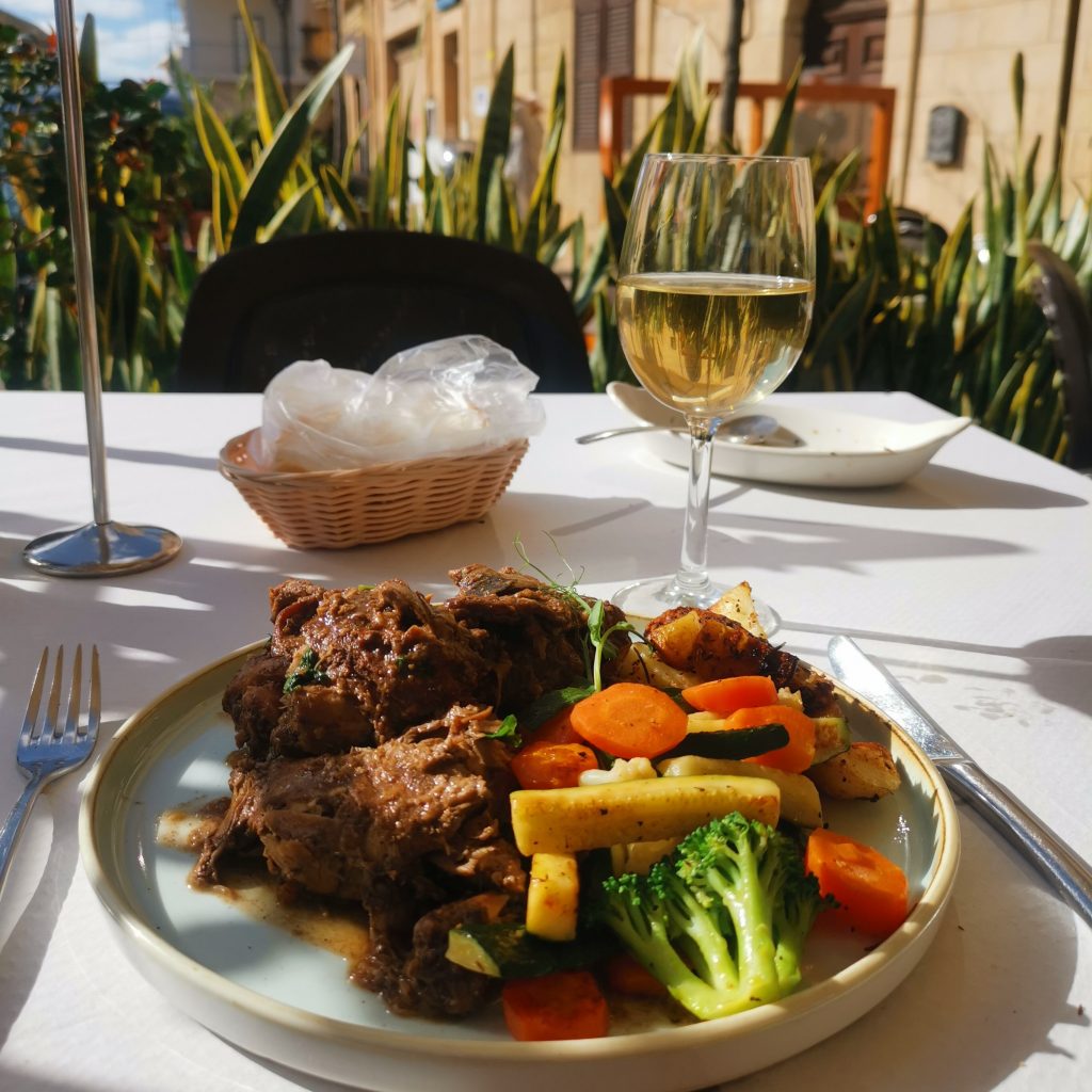 Plate of rabbit stew, roast potatoes, and seasonal vegetables on a plate on restaurant table with glass of white wine 