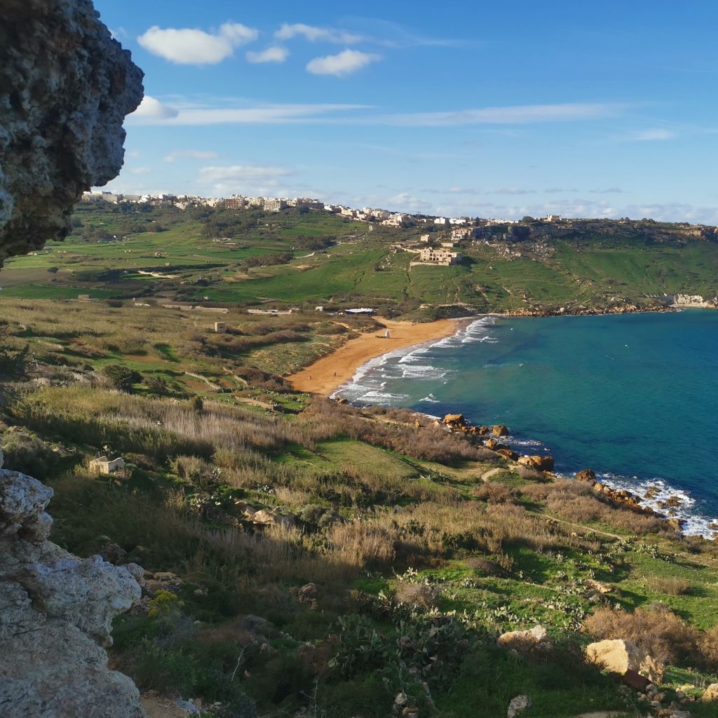 Hiking routes in Gozo title image - iew of orange sand and blue ocean from above with green hills in background and grassy areas and brush in foreground