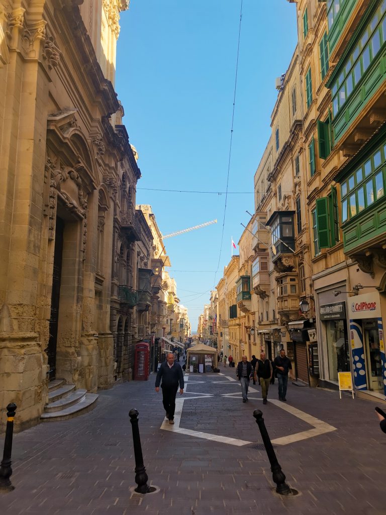 View of a street in Valetta with tall beige buildings with green windowed balconies